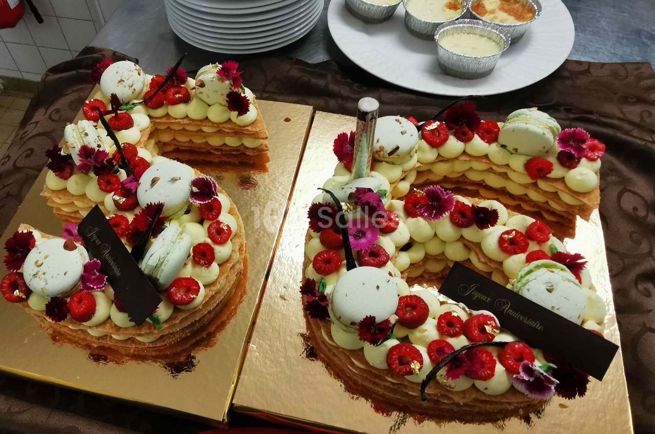 Gâteau en forme de chiffre décoré de fleurs comestibles, macarons et fruits rouges, posé sur un plateau doré.