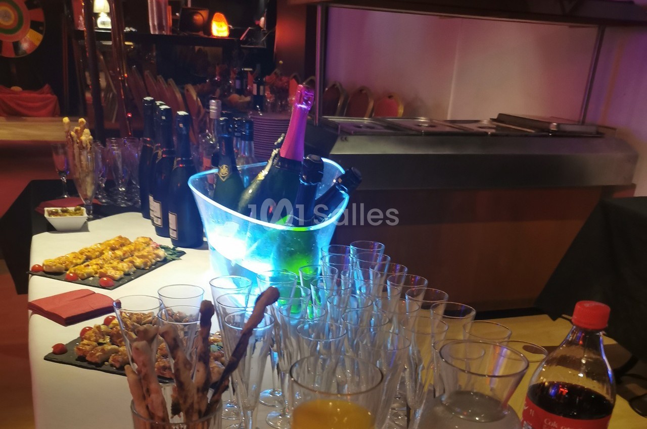 Table dressée avec des verres, une bouteille de champagne dans un seau lumineux, amuse-bouches et boissons variées.