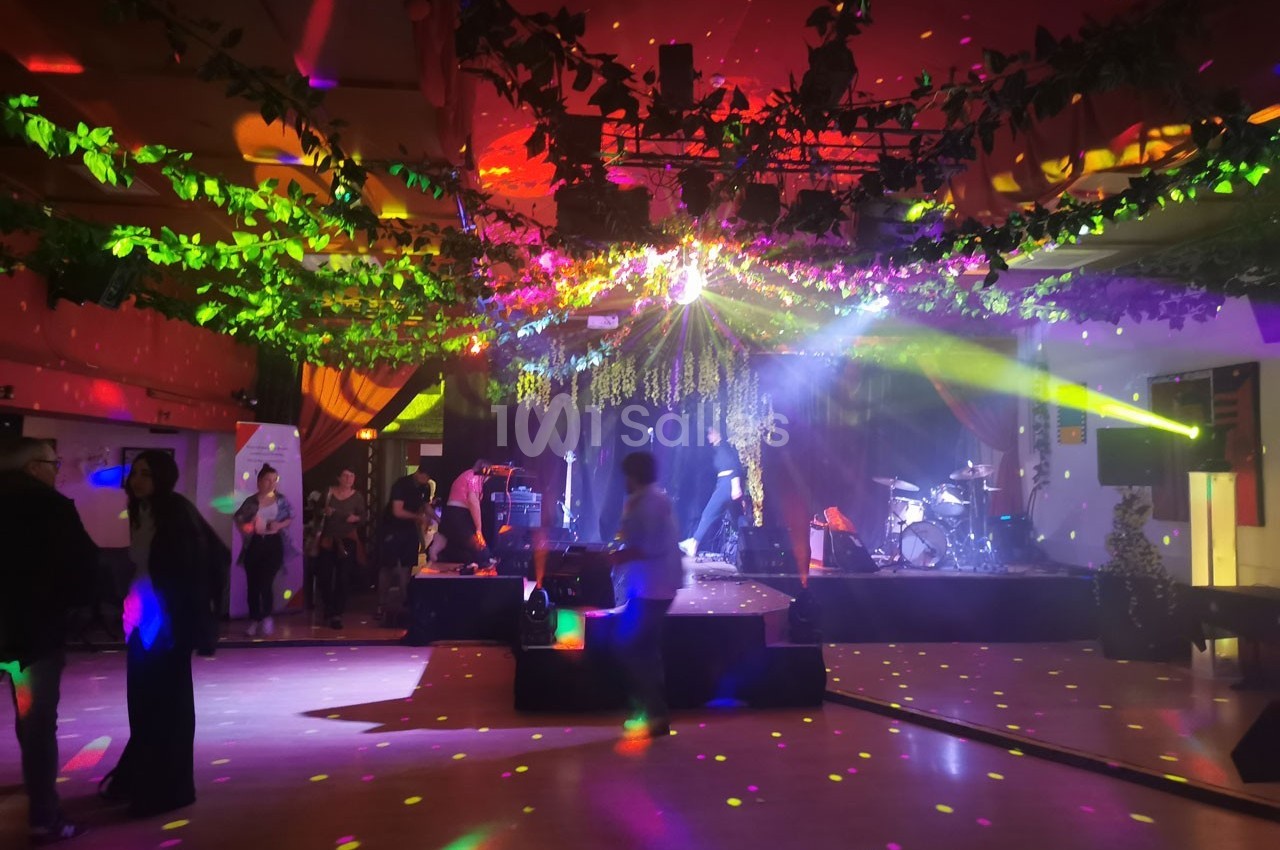 Salle de fête décorée de guirlandes lumineuses et végétales, avec une scène, des instruments et des jeux de lumière colorés.