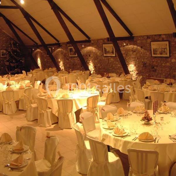 Salle de réception avec des tables rondes dressées, nappes blanches, chaises décorées et murs en pierre apparente.