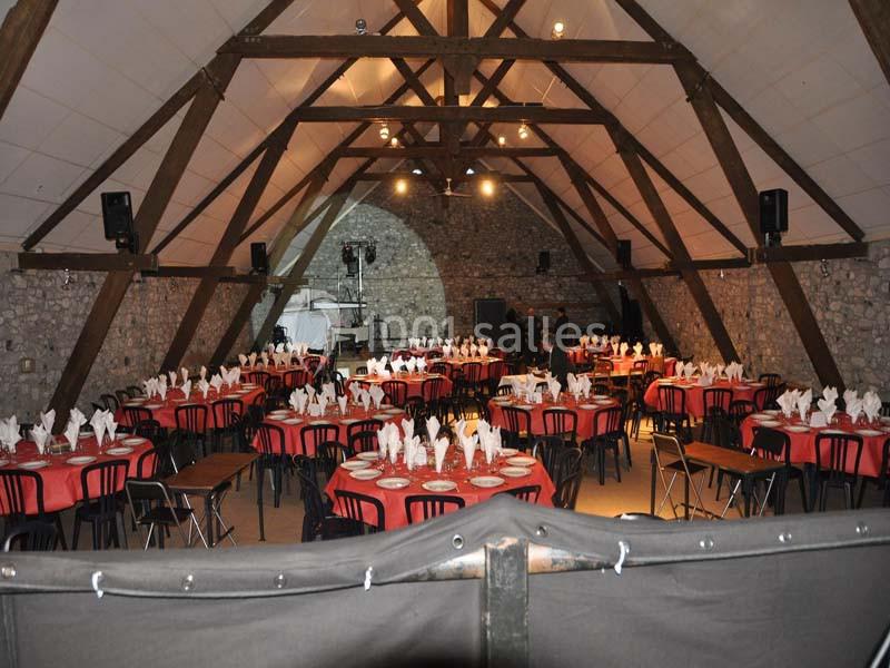 Salle en pierre avec charpente apparente, tables rondes dressées avec nappes rouges et serviettes pliées.