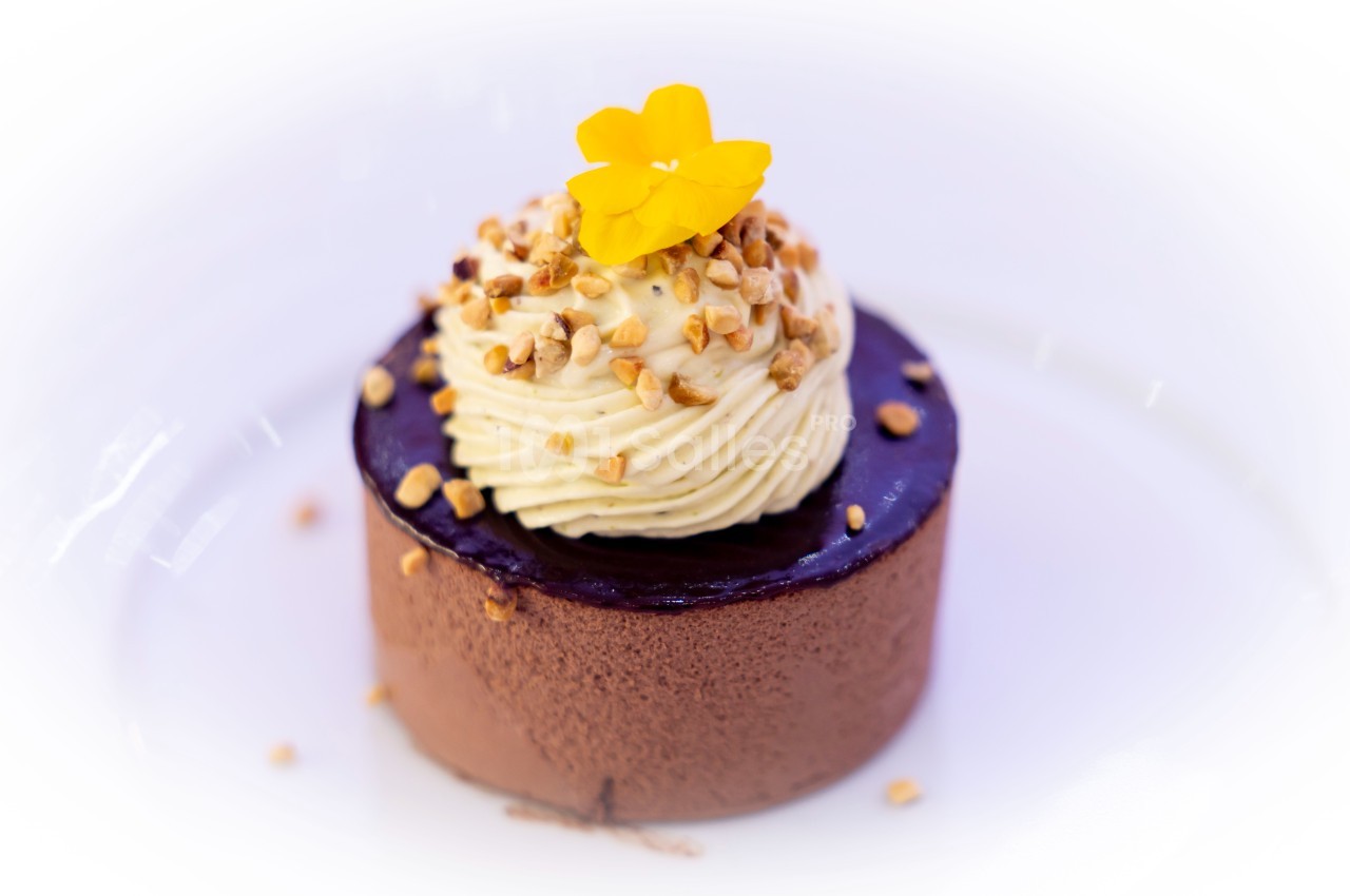 Dessert au chocolat avec glaçage brillant, crème fouettée, éclats de noisettes et fleur jaune décorative.