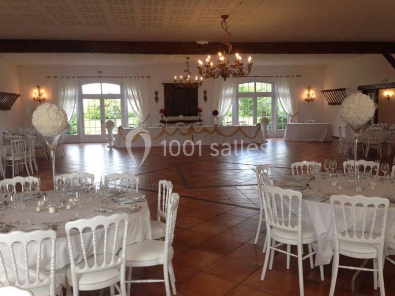 Salle de réception lumineuse avec tables rondes dressées, chaises blanches et décorations florales élégantes.