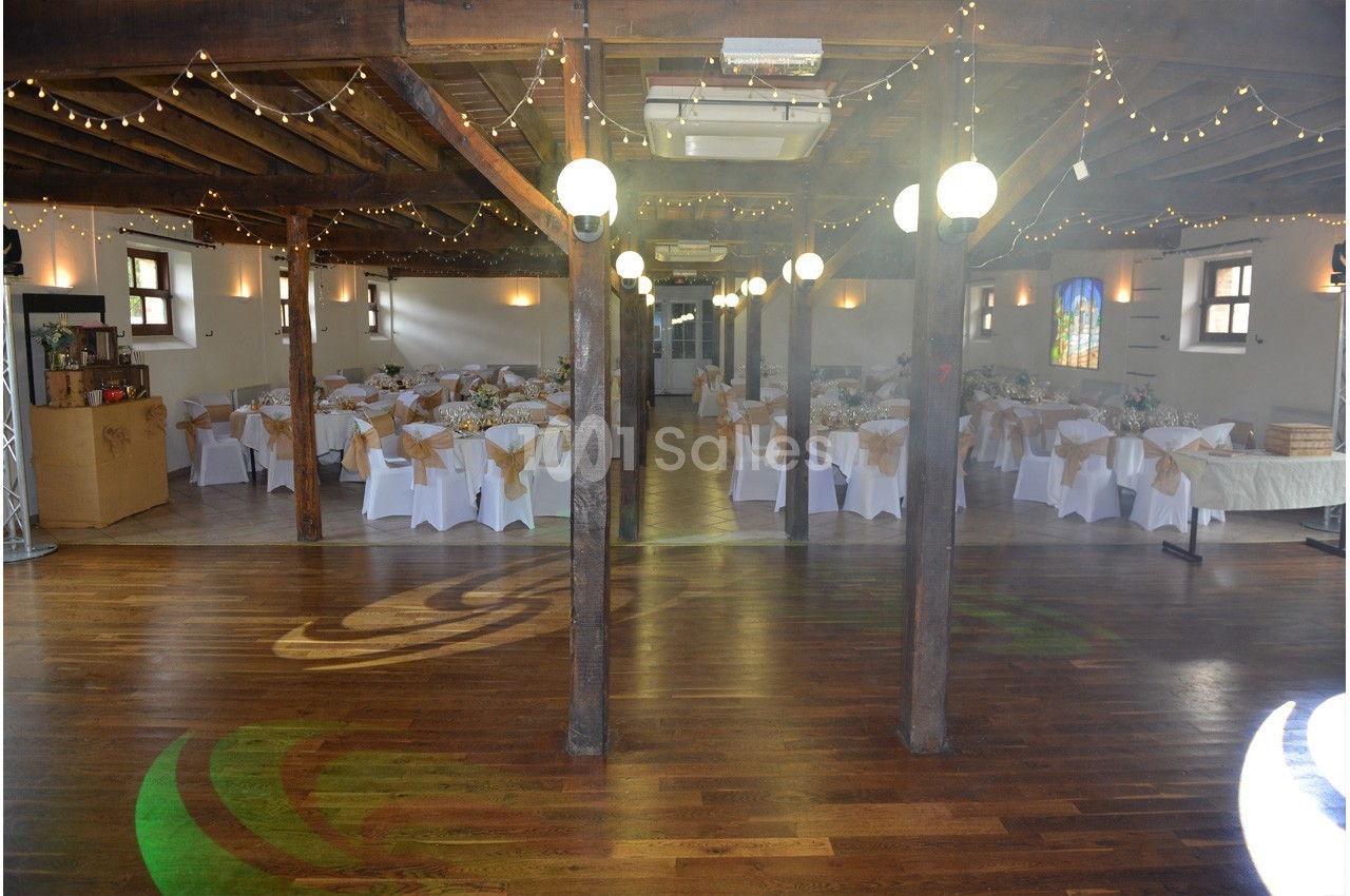 Salle de réception avec tables décorées de nappes blanches et chaises, éclairée par des guirlandes lumineuses suspendues.