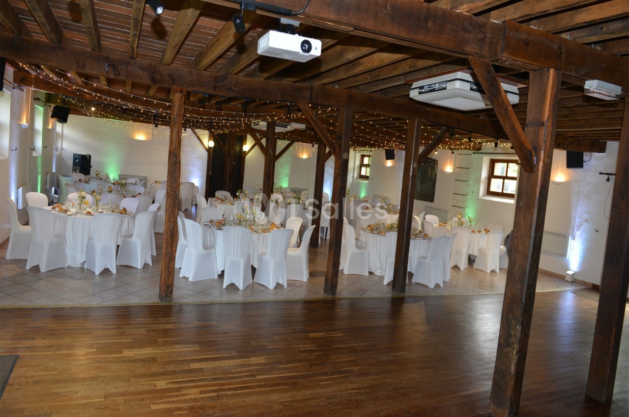 Salle de réception avec tables rondes dressées, chaises blanches et décorations lumineuses sous un plafond en bois.