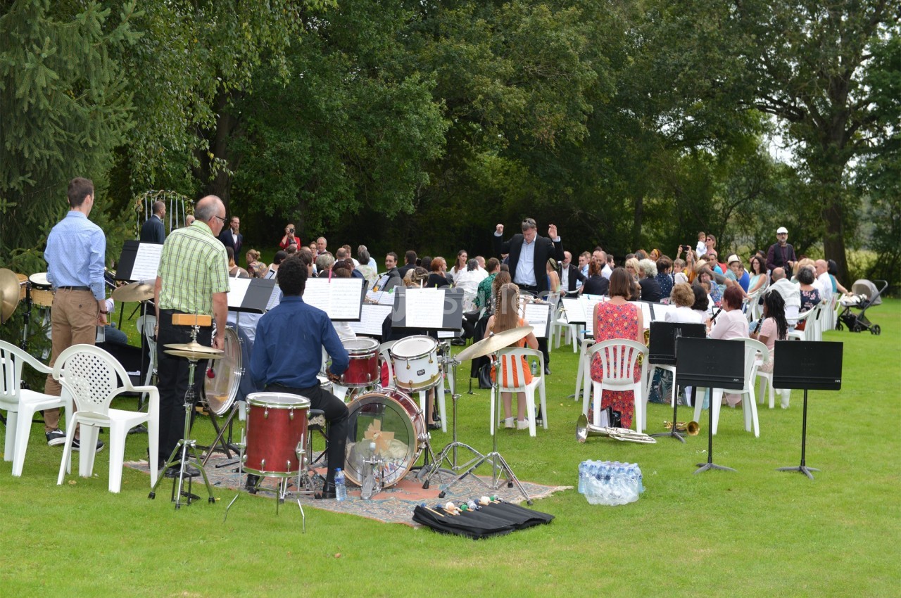 Un orchestre joue en plein air devant un public assis sur des chaises blanches dans un parc verdoyant.