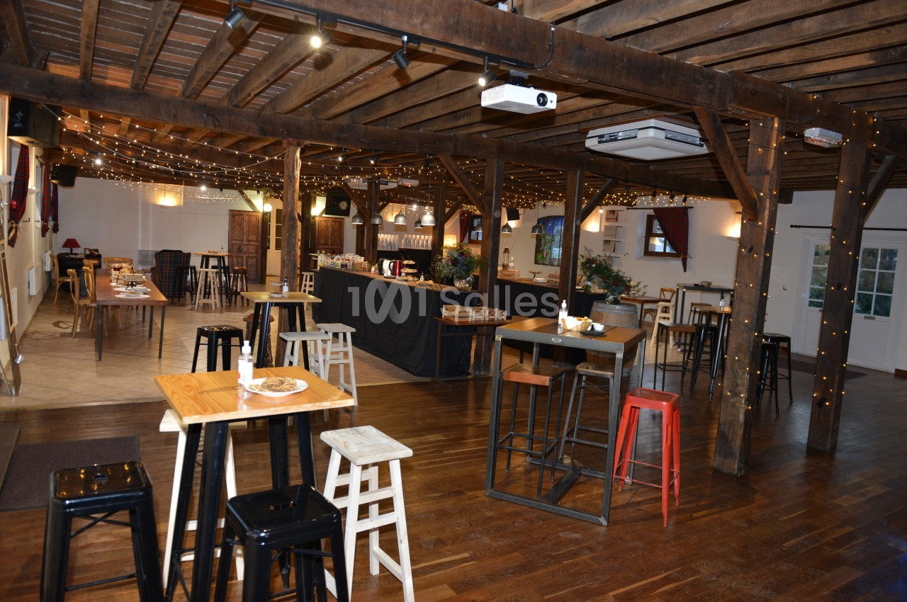 Salle en bois avec tables hautes, chaises variées, guirlandes lumineuses et comptoir en arrière-plan.