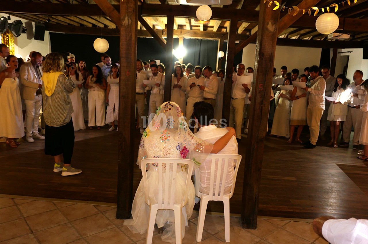 Un couple assis de dos lors d'une fête, entouré d'invités vêtus de blanc dans une salle décorée.
