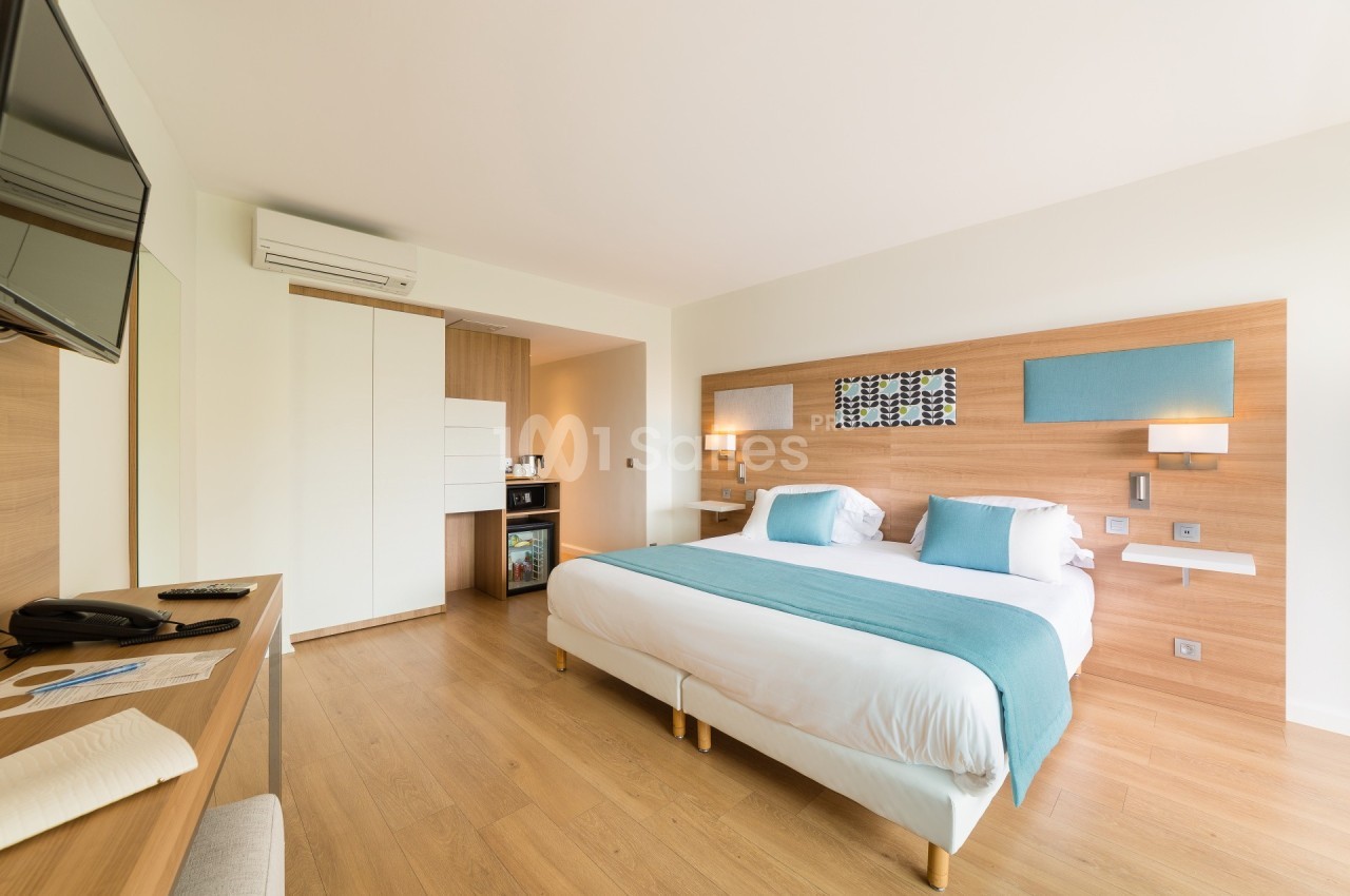Chambre lumineuse avec lit double, décor moderne en bois clair, télévision murale et coin minibar.