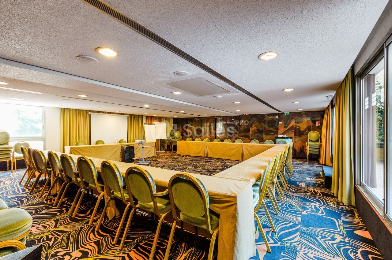 Salle de réunion avec tables disposées en U, chaises vertes, tapis coloré et grandes fenêtres laissant entrer la lumière…