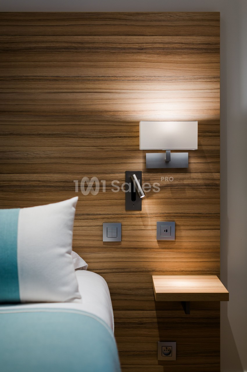 Lampe murale allumée au-dessus d'une table de chevet en bois, près d'un lit avec une housse turquoise et blanche.