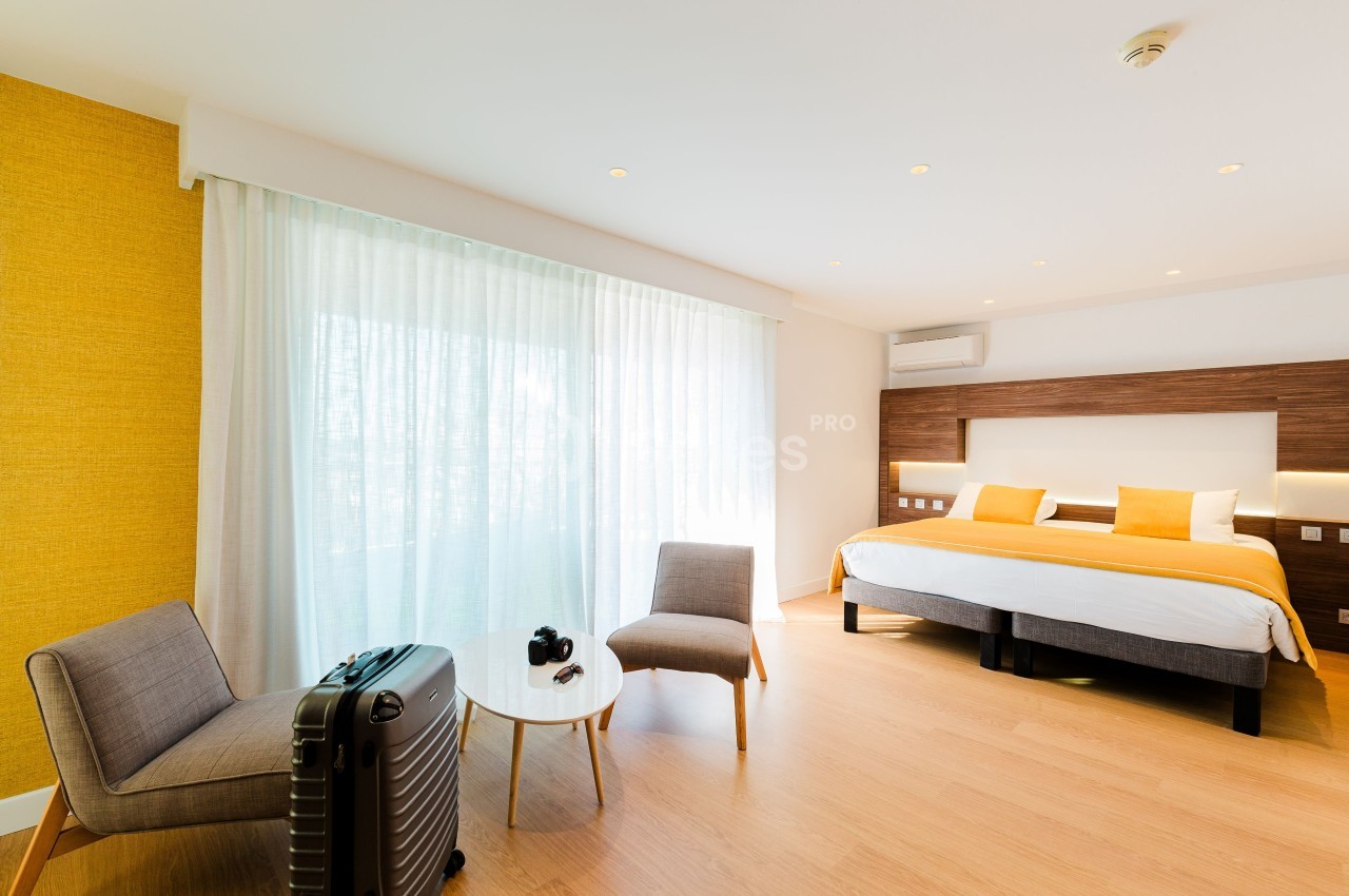 Chambre lumineuse avec lit double, fauteuils, table basse, valise et rideaux blancs laissant entrer la lumière naturelle.