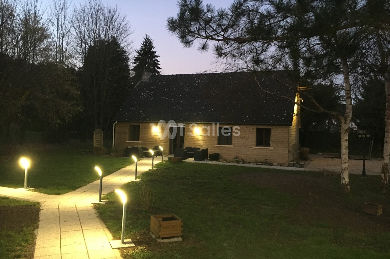 Maison en bois éclairée à la tombée de la nuit, entourée d'arbres et d'un chemin bordé de lampadaires.