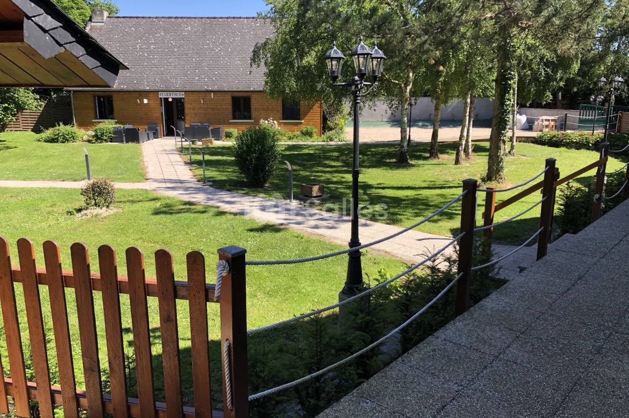 Jardin verdoyant avec allée pavée, clôture en bois, lampadaire et bâtiment en pierre sous un ciel dégagé.