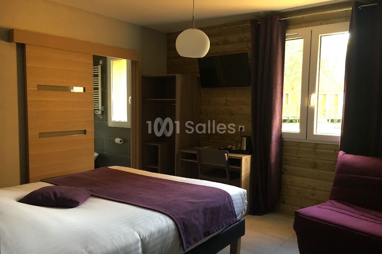Chambre lumineuse avec lit double, bureau, télévision murale, fauteuil et fenêtre donnant sur l'extérieur.