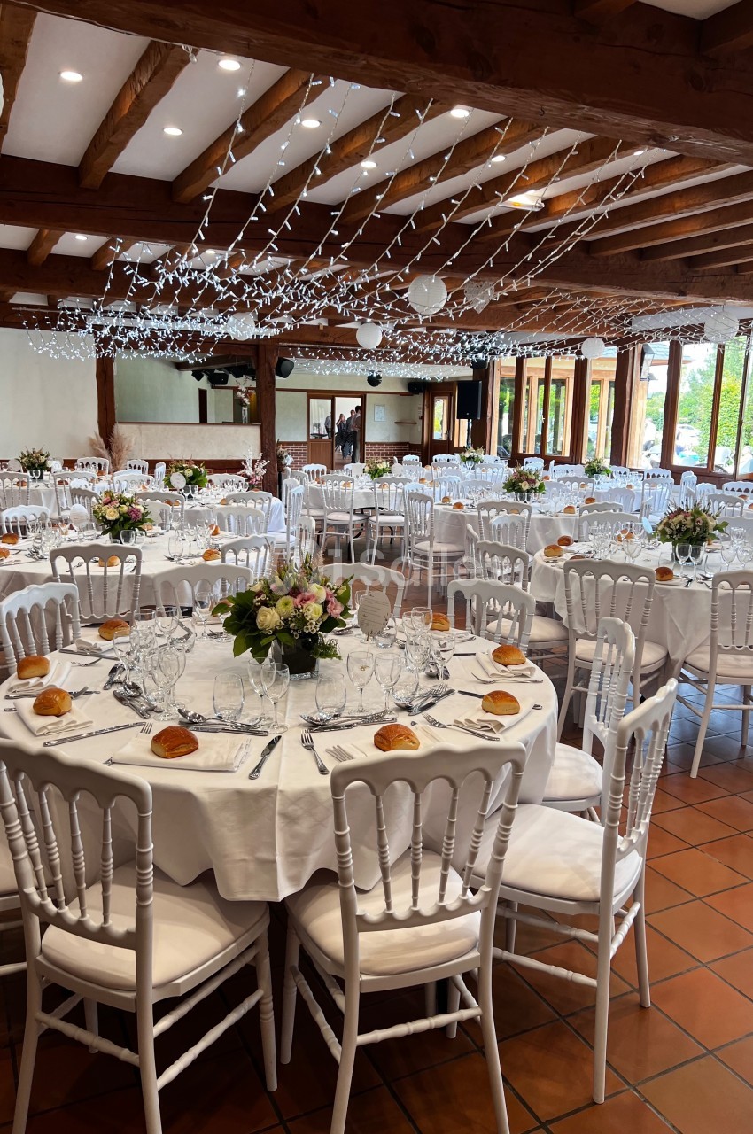 Salle de réception décorée avec des tables rondes dressées, des chaises blanches et des guirlandes lumineuses suspendues.