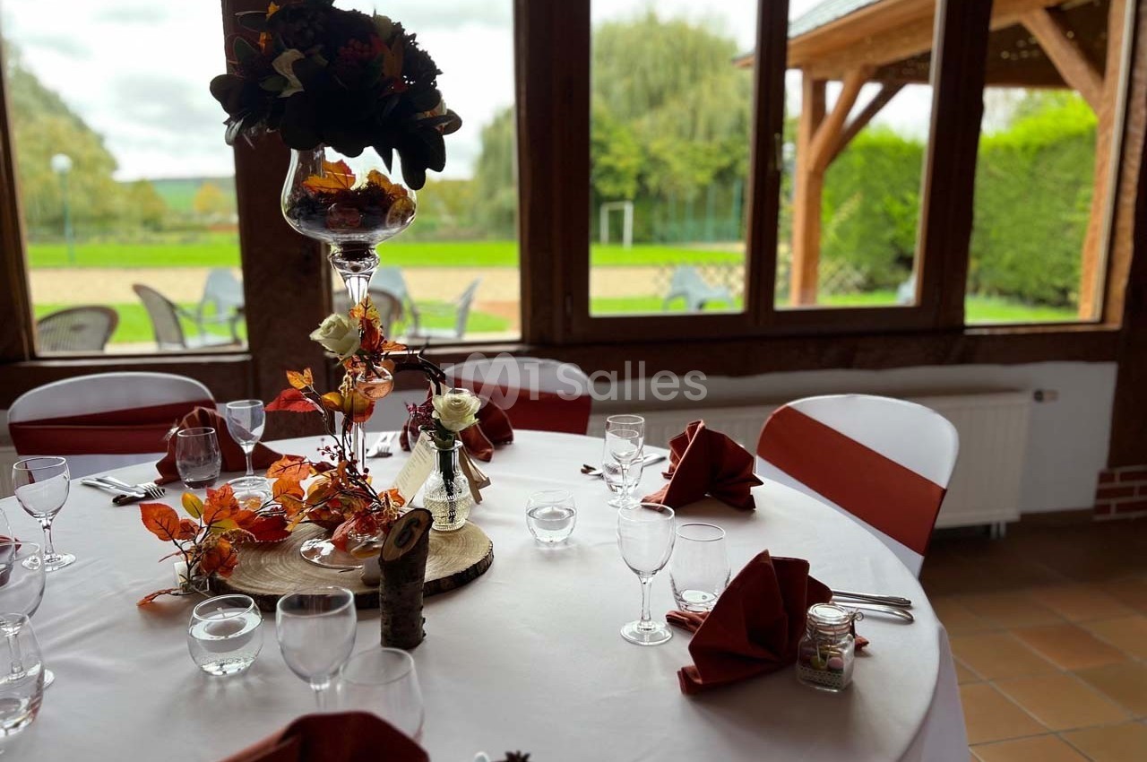 Table ronde décorée pour un événement, ornée de fleurs automnales, nappes blanches et serviettes rouges pliées.