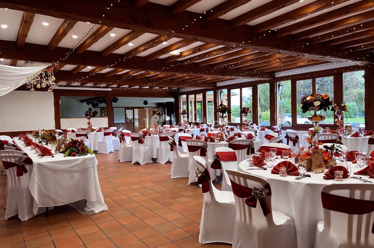 Salle de réception décorée pour un événement, avec tables rondes dressées, chaises ornées de rubans rouges et fleurs…