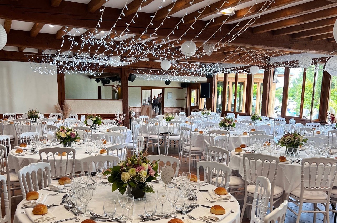Salle de réception décorée pour un événement, avec tables rondes dressées, fleurs et guirlandes lumineuses suspendues.