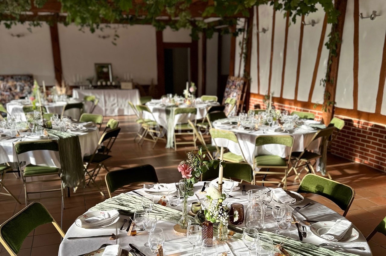 Salle de réception décorée avec des tables rondes dressées, nappes blanches, chaises vertes et feuillage suspendu.
