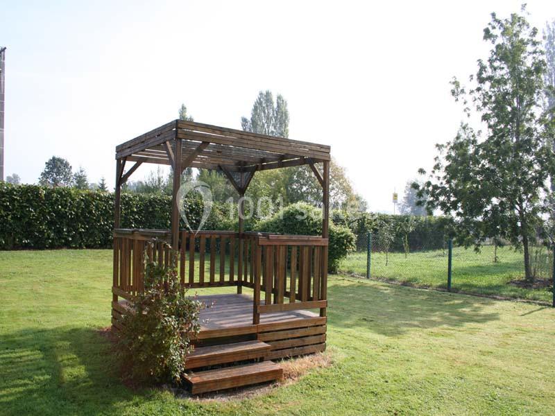 Tonnelle en bois avec escalier, située sur une pelouse entourée de haies et d'arbres dans un jardin.
