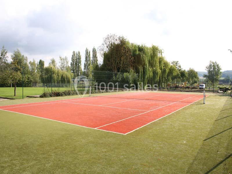 Terrain de tennis extérieur en gazon synthétique rouge, entouré d'arbres et de verdure sous un ciel partiellement nuageux.