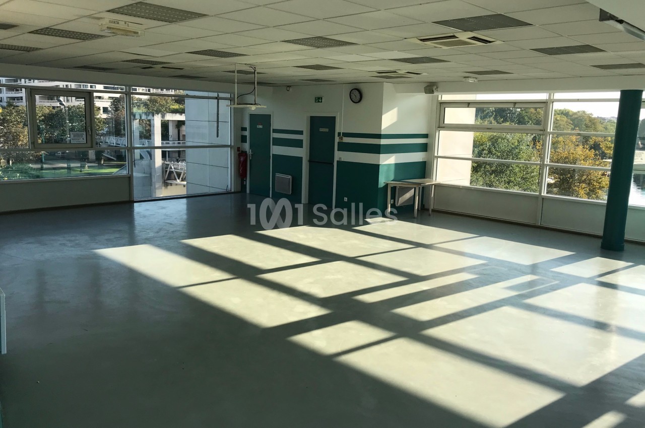 Salle vide avec sol en béton, grandes fenêtres laissant entrer la lumière naturelle et vue sur des arbres à l'extérieur.