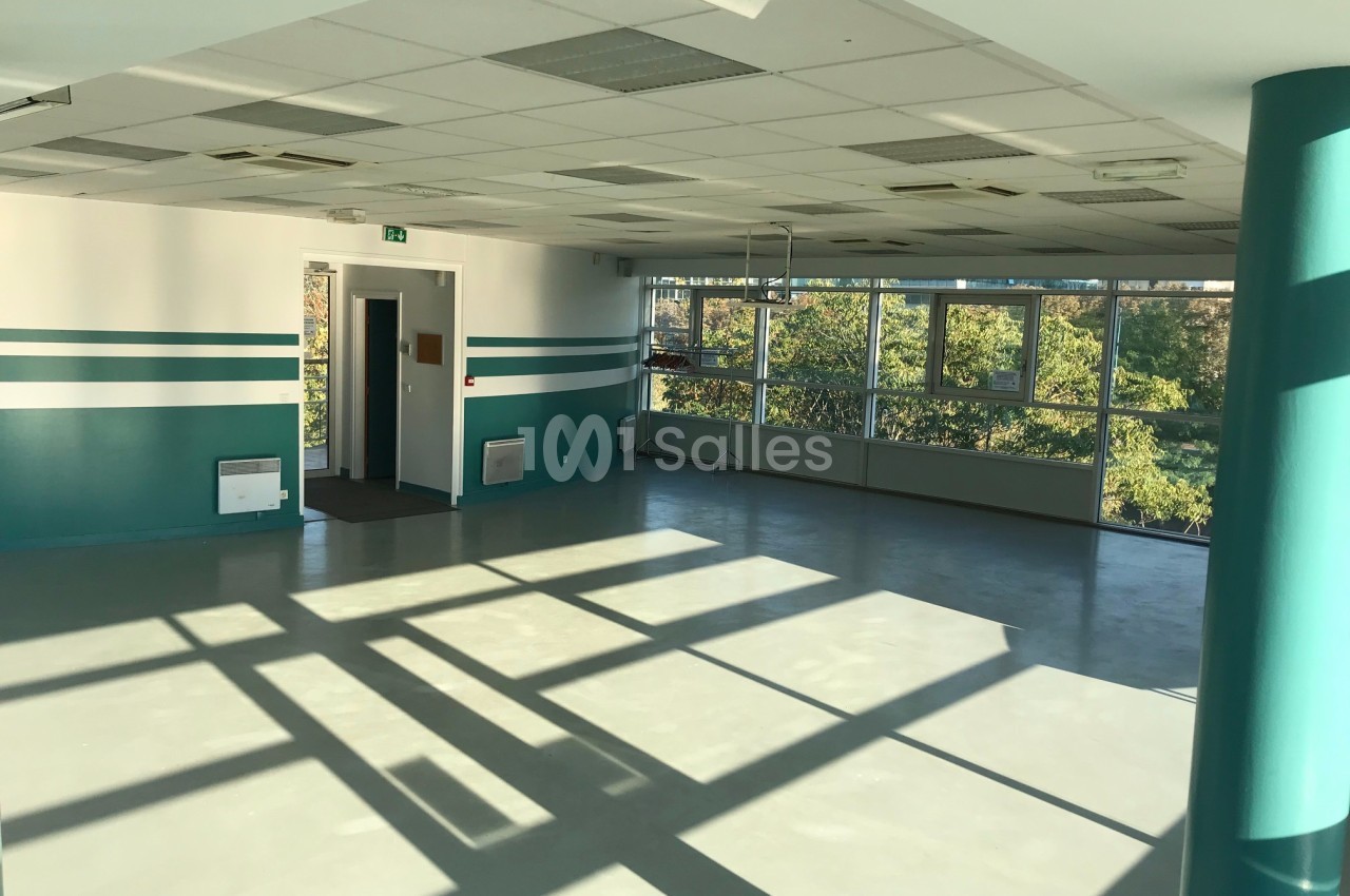 Salle vide avec sol en béton, murs peints en blanc et vert, grandes fenêtres laissant entrer la lumière naturelle.