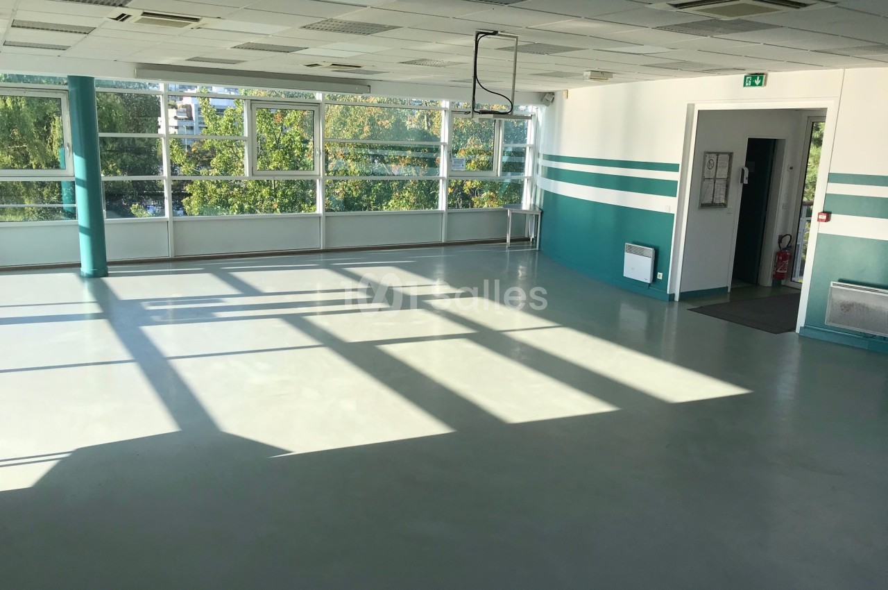 Salle vide lumineuse avec sol clair, grandes fenêtres laissant entrer la lumière naturelle et murs blancs et verts.