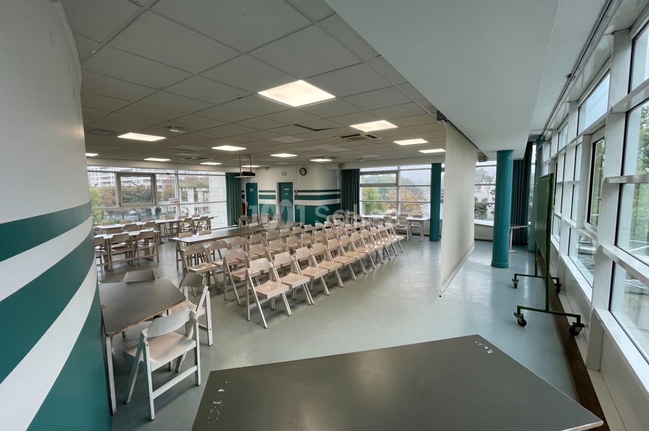 Salle lumineuse avec rangées de chaises blanches, tables et écran de projection, entourée de grandes fenêtres.