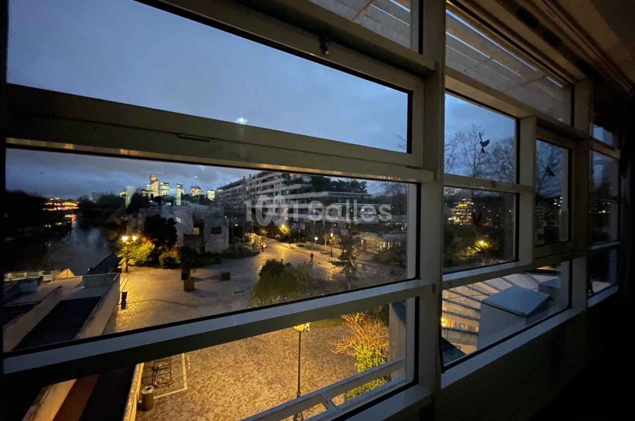 Vue nocturne d'une place éclairée avec des bâtiments modernes et un cours d'eau, observée depuis une fenêtre.
