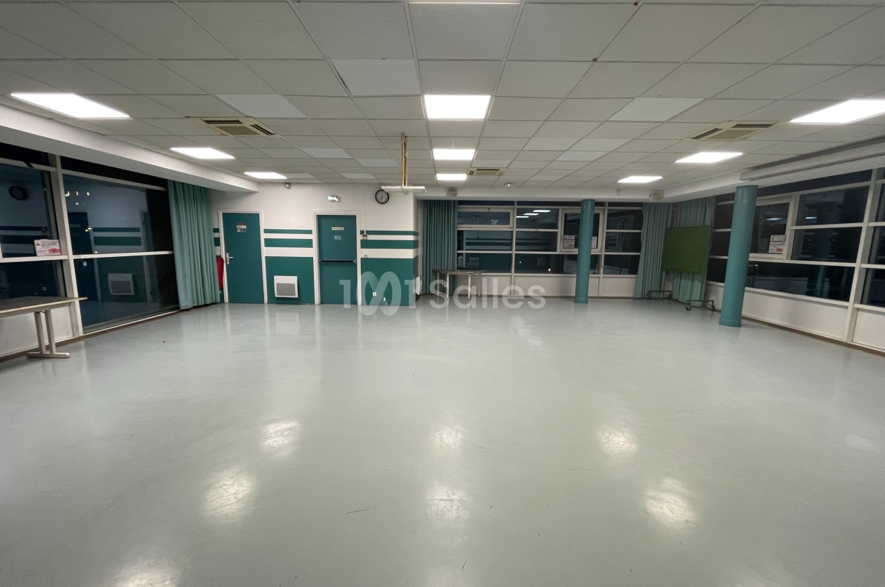 Salle vide avec sol clair, murs verts et blancs, grandes fenêtres, éclairage au plafond et mobilier minimal.