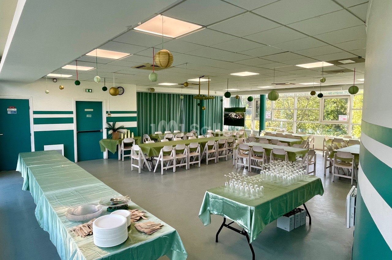 Salle décorée pour un événement, avec tables dressées, nappes vertes, chaises blanches et lanternes suspendues.