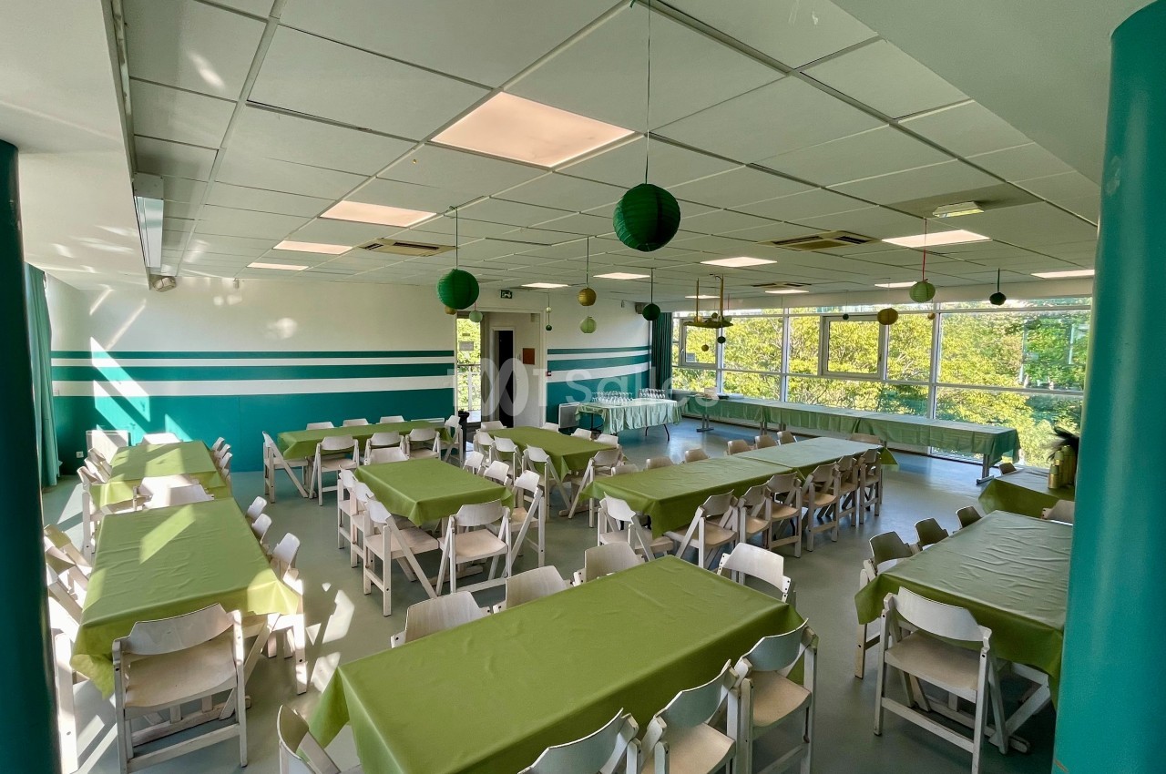 Salle lumineuse avec des tables recouvertes de nappes vertes, chaises blanches et décorations suspendues.