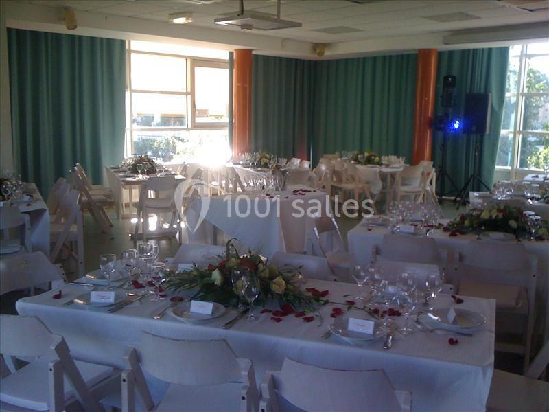 Salle décorée pour un événement, avec tables dressées, nappes blanches, fleurs et chaises pliantes blanches.