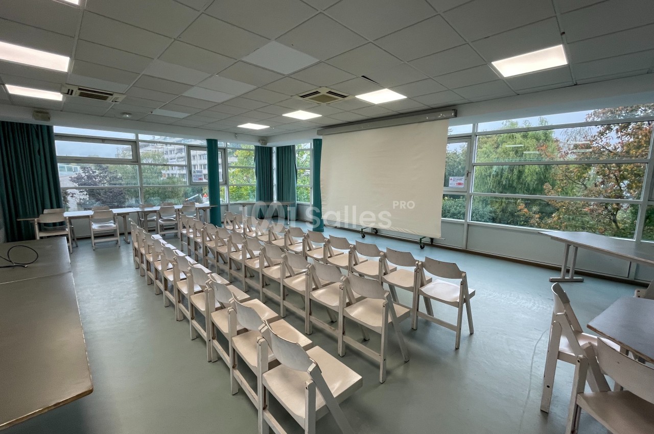 Salle de conférence lumineuse avec des rangées de chaises blanches, un écran de projection et de grandes fenêtres donnant…