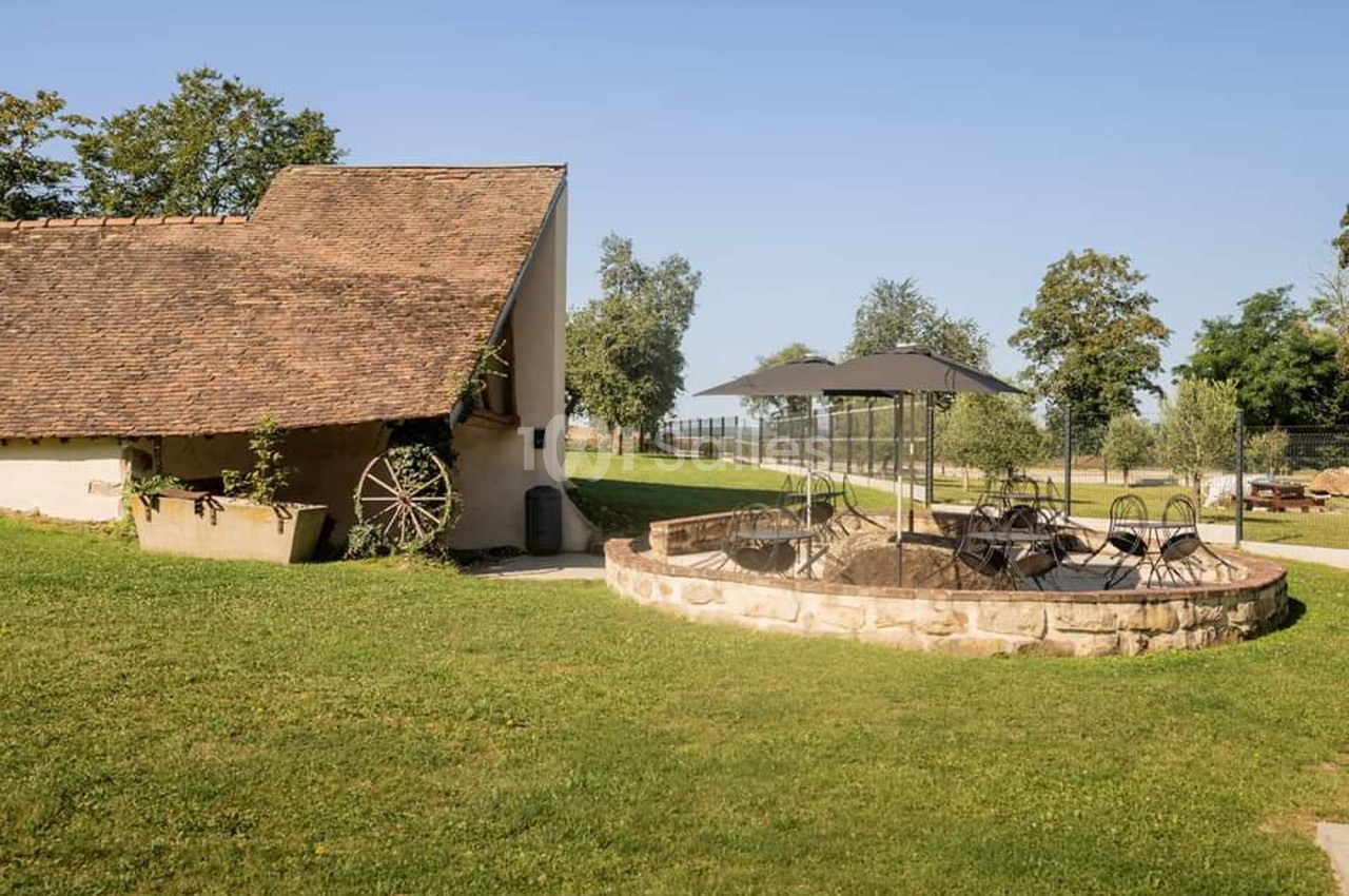 Cour extérieure avec pelouse, terrasse en pierre, mobilier de jardin sous un parasol et bâtiment ancien en arrière-plan.