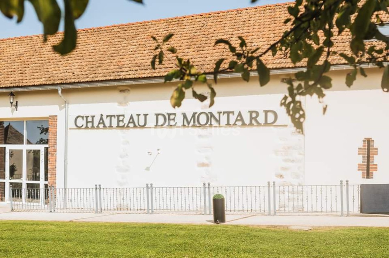 Façade d'un bâtiment avec l'inscription ’Château de Montard’, entourée de pelouse et partiellement ombragée.
