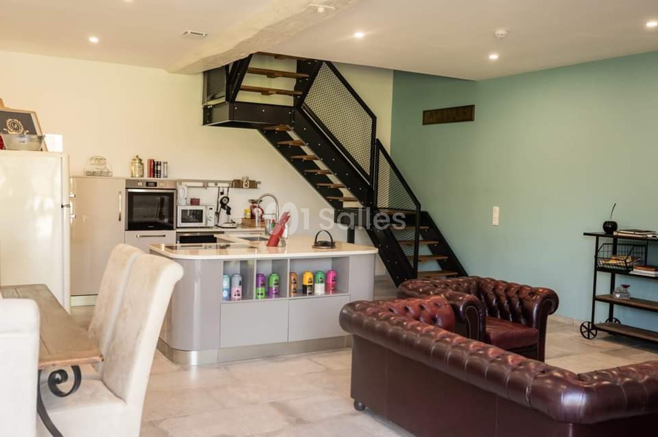 Cuisine ouverte moderne avec îlot central, canapé en cuir, escalier métallique et mur bleu en arrière-plan.