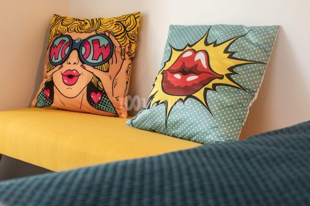 Deux coussins colorés au style pop art posés sur un banc jaune, dans un intérieur lumineux.