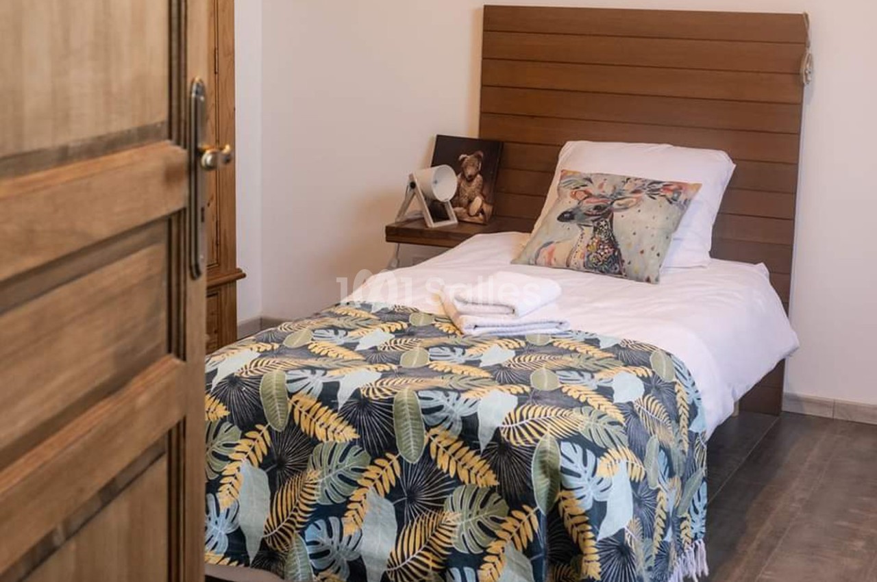 Lit simple avec une tête de lit en bois, couvert d'une couverture à motifs tropicaux, dans une chambre lumineuse.