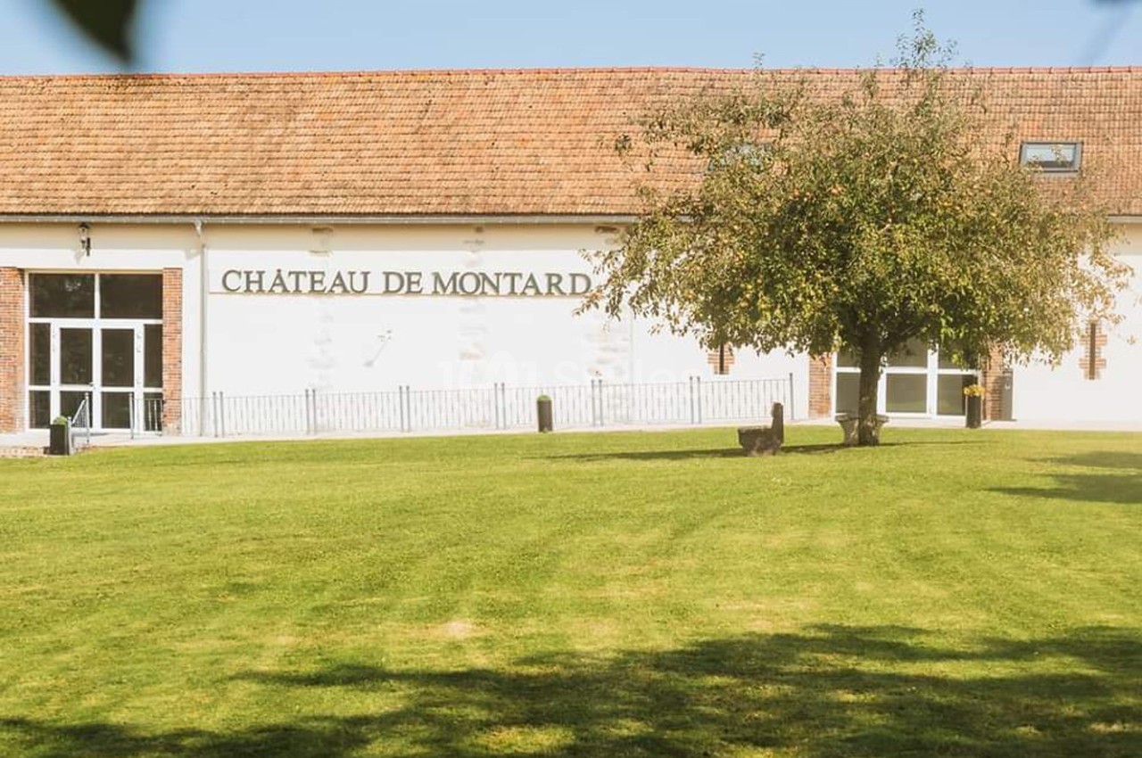 Bâtiment blanc avec toit en tuiles rouges, marqué ’Château de Montard’, entouré d'une pelouse et d'un arbre.