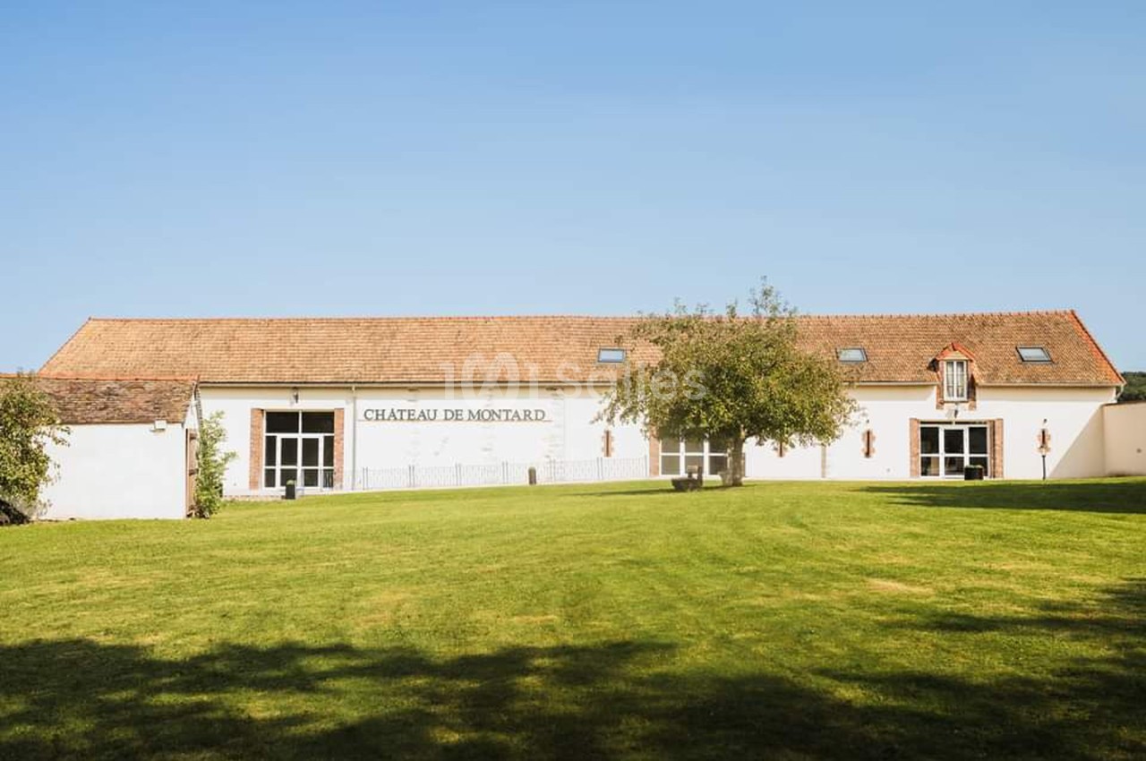 Bâtiment blanc avec toit en tuiles rouges, entouré d'une pelouse et d'arbres sous un ciel dégagé.