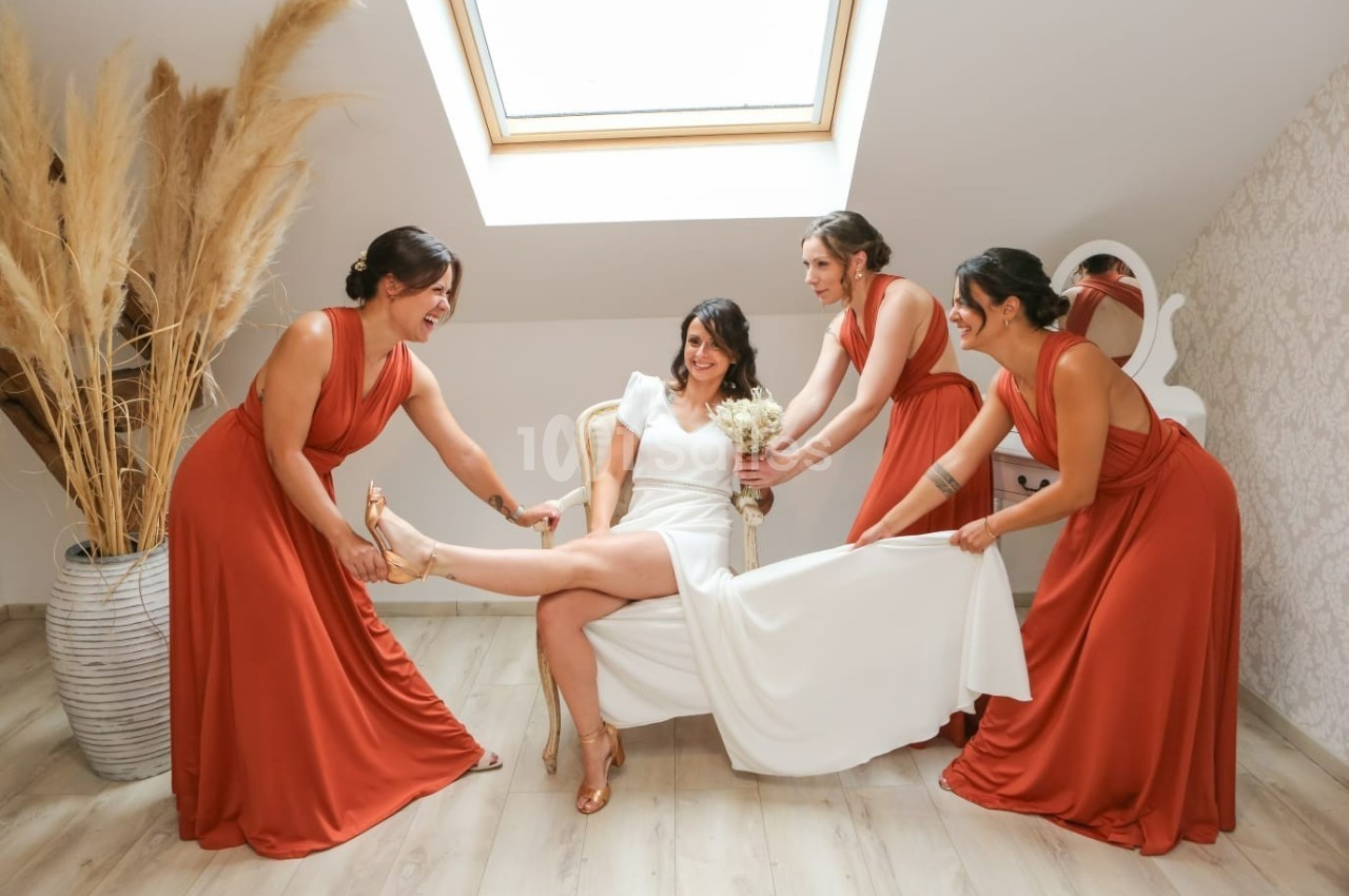 Une mariée souriante en robe blanche est entourée de trois femmes en robes orange ajustant ses chaussures.