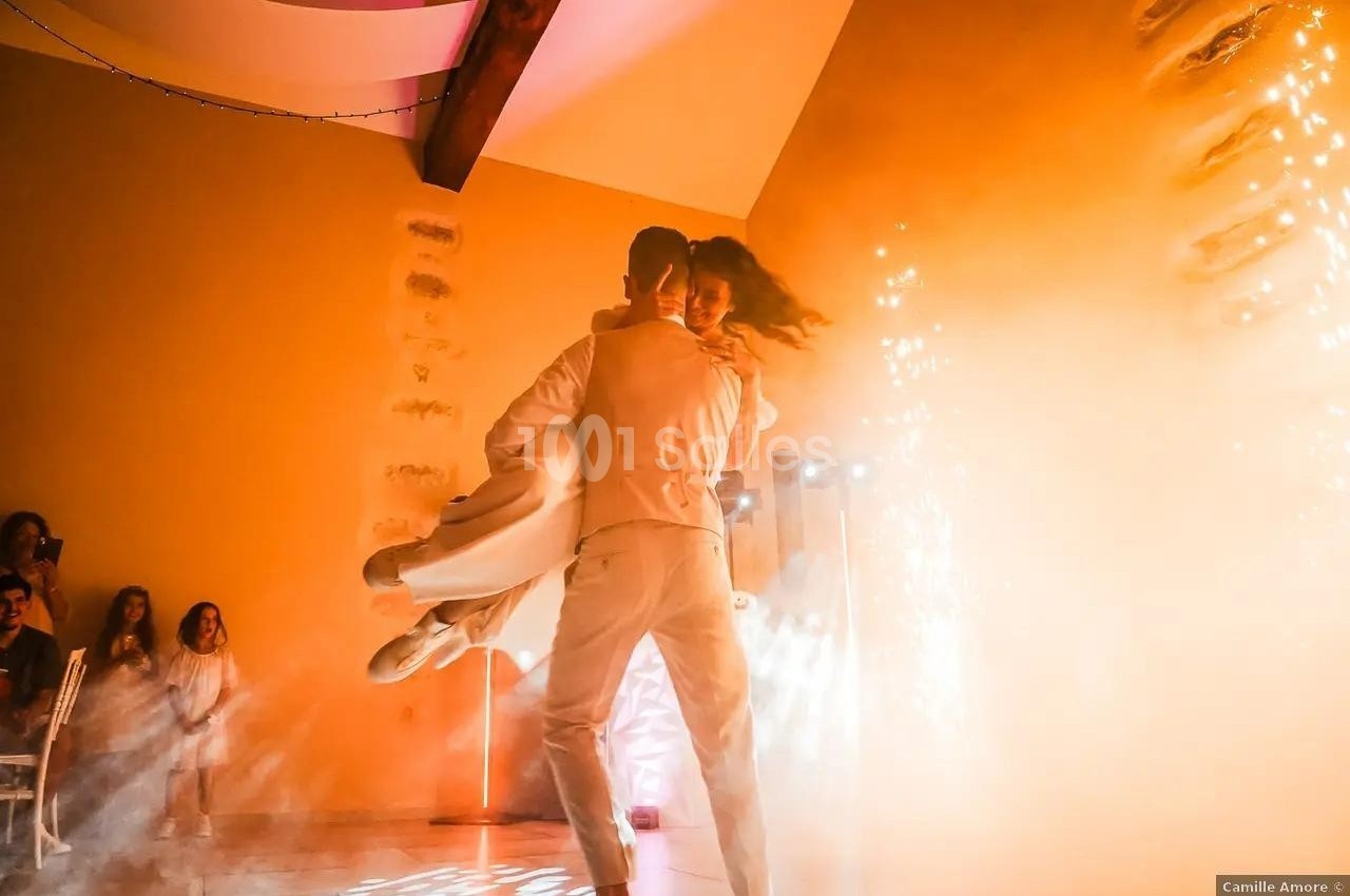 Un couple danse dans une salle illuminée, entouré de fumée et d'étincelles, sous le regard des invités.