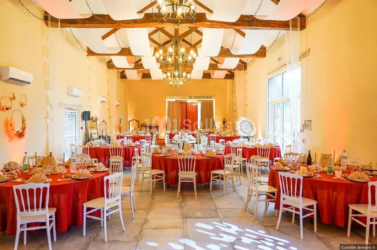Salle de réception décorée avec des tables rondes, nappes rouges, chaises blanches et plafond avec poutres apparentes.