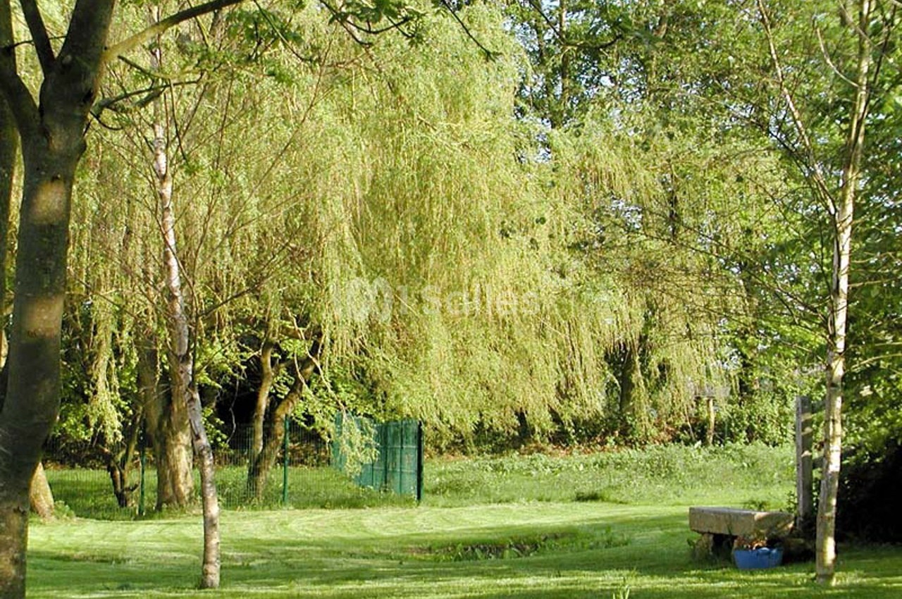 Un jardin verdoyant avec des arbres, une pelouse tondue, un banc en pierre et une clôture en arrière-plan.