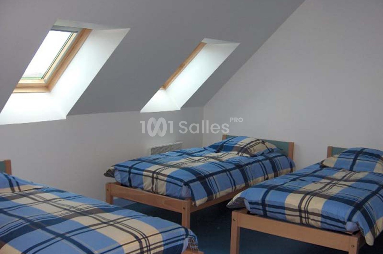 Trois lits simples avec des couvertures à carreaux bleus dans une chambre mansardée avec fenêtres de toit.
