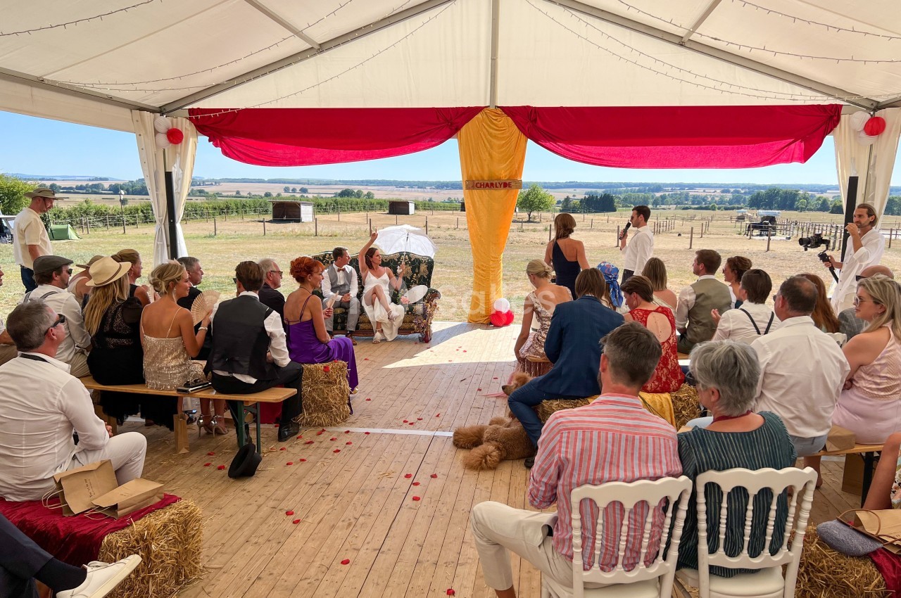 Cérémonie en plein air sous une tente décorée, avec des invités assis sur des bottes de paille et des chaises.