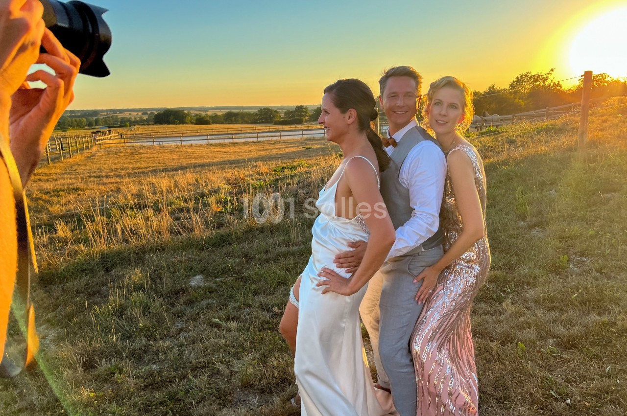 Trois personnes posent en tenue élégante dans un champ au coucher du soleil, avec un photographe au premier plan.